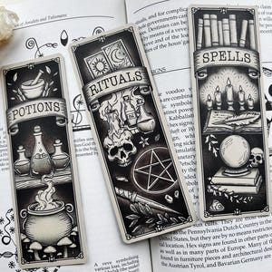 May include: Three black and white illustrated bookmarks with occult themes. One says "Potions" with a cauldron, another "Rituals" with a skull, and the third "Spells" with candles. The bookmarks are on top of an open book.
