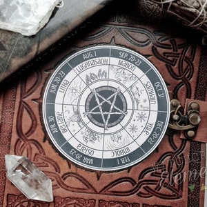 May include: A white circle with a black pentacle in the center and a wheel of the year with the names of the eight sabbats and their dates. The circle is on a brown leather journal with a clasp.