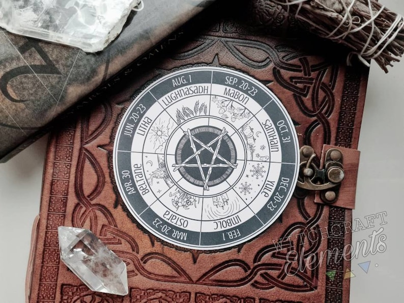 May include: A white circle with a black pentacle in the center and a wheel of the year with the names of the eight sabbats and their dates. The circle is on a brown leather journal with a clasp.