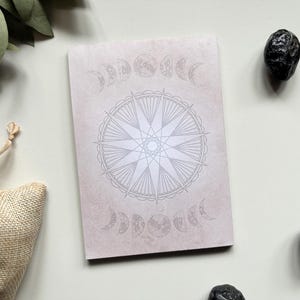 May include: A beige notebook with a celestial design featuring a compass rose and moon phases. The notebook is surrounded by black stones and a small hessian bag.