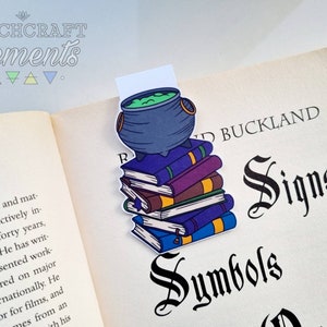 May include: A bookmark depicting a stack of books topped with a cauldron containing green liquid. The bookmark is inserted into a book titled "Signs Symbols". The image also shows the text "Witchcraft Elements" at the top.