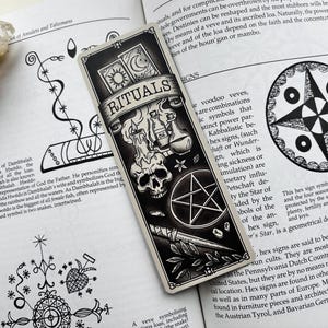 May include: Black and white bookmark with the word "RITUALS" on a banner. The bookmark features illustrations of a skull, pentagram, candles, and other occult symbols. The bookmark is resting on an open book.