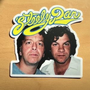 May include: A sticker featuring a photograph of two men with the text "Steely Dan" in yellow script with a blue outline. The men are facing forward, with dark hair and light skin.