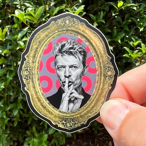 May include: A sticker featuring a portrait of David Bowie within a gold-colored ornate frame. The background behind the portrait is gray with red circular patterns. Bowie is depicted with his finger to his lips, in a gesture of silence.