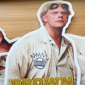May include: A sticker featuring a character with goggles and a lab coat. The sticker has the word "BROWN" in gold letters at the bottom. The character has a surprised expression. The sticker is white with a white border.