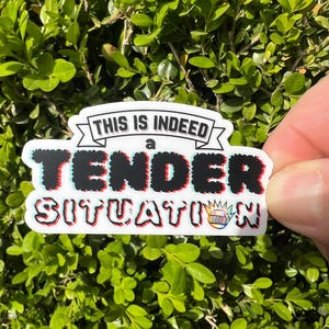 May include: A white sticker with the text "THIS IS INDEED a TENDER SITUATION" in black, red, and blue. The sticker has a banner design and a small crown graphic. The sticker is held against a background of green leaves.