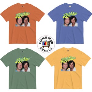 Könnte beinhalten: Vier kurzärmelige T-Shirts mit Rundhalsausschnitt in Orange, Blau, Grün und Gelb. Jedes Shirt zeigt einen Grafikdruck von zwei Männern mit dem Schriftzug Steely Dan in gelber Schreibschrift darüber. Ein kreisförmiges Logo von Couch Tour Sticker Co. ist in der Mitte.