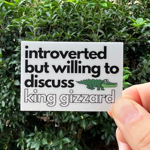 May include: White sticker with black text that reads "introverted but willing to discuss king gizzard." A green cartoon alligator is featured in the design. The sticker is held in front of a green leafy background.