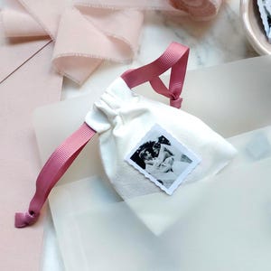 Puede incluir: Una pequeña bolsa de tela de color blanco roto con cordón, que presenta una fotografía cuadrada en blanco y negro con borde festoneado en la parte frontal. La bolsa se cierra con dos cintas de grosgrain rosa empolvado.