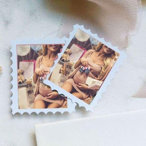 Puede incluir: Dos fotos en forma de sello postal azul claro de una persona embarazada sosteniendo zapatos de bebé. Las fotos están sobre una superficie blanca con una ramita de flores secas y una cinta rosa. Las fotos están rasgadas.