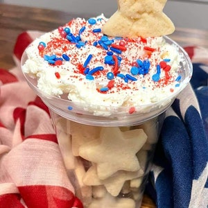 May include: A clear plastic cup filled with star-shaped sugar cookies topped with whipped cream and red, white and blue sprinkles. A star-shaped cookie sits on top of the whipped cream.