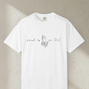 May include: White t-shirt with a crew neck. The shirt features a black and white graphic of praying hands with the text "perdi la en D.C." printed above the hands.