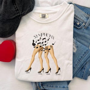 Puede incluir: Camiseta blanca con la palabra "SYMPHONY" y notas musicales sobre tres pares de piernas con tacones altos. Las palabras "CLICK" y "CLACK" est&aacute;n impresas debajo. Una gorra negra, un bolso rojo en forma de coraz&oacute;n y unos vaqueros tambi&eacute;n est&aacute;n en la imagen.