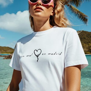 Könnte beinhalten: Weißes T-Shirt mit dem Text "un mal en madrid" und einer herzförmigen Ballon-Grafik. Die Person trägt eine rosa Sonnenbrille. Der Hintergrund umfasst eine Palme, blauen Himmel und Ozean.