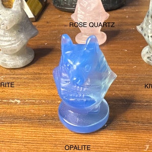 May include: A collection of carved crystal figurines, including Labradorite, Rose Quartz, Opalite, and Kiwi Jasper. Each figurine is a stylized cat head on a pedestal base. The background includes a black statue and other crystals.