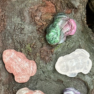 May include: Five carved gemstone frog figurines in various colors, including pink, green, and purple. The frogs are displayed on a textured, dark brown surface, showcasing their unique shapes and colors.