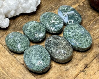 May include: A collection of eight polished green moss agate stones, each with unique patterns and textures. The stones are oval-shaped and rest on a wooden surface, showcasing their natural beauty. The stones vary in shades of green and white.