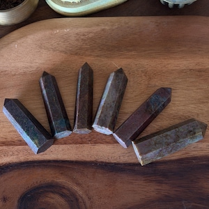 May include: A collection of decorative items on a wooden surface. The items include six dark brown, faceted crystal points, a small bronze bowl, a light green figurine, and two ornate metal containers.