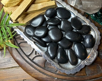 May include: A collection of natural elements, including smooth, black stones in a silver dish, several pieces of Palo Santo wood, and large clear quartz crystals. The items are arranged on a wooden surface, creating a natural and earthy aesthetic.