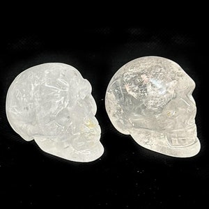 May include: Three clear quartz crystal skull carvings. Each skull is carved with detailed features, including eye sockets, a nose cavity, and teeth. The skulls are displayed against a black background.