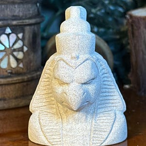 May include: A light gray stone sculpture of a bird-headed figure, possibly Egyptian. The sculpture has a detailed face with closed eyes and a layered headdress. The figure is seated and has a textured surface.