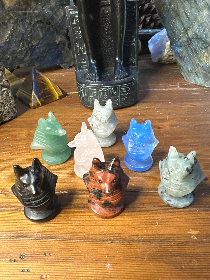 May include: A collection of carved stone wolf figurines in various colors, including black, red, green, and blue. The figurines are displayed on a wooden surface, with an Egyptian statue in the background. The wolf figurines are a unique decorative item.