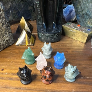 May include: A collection of carved gemstone wolf figurines in various colors, including green, pink, blue, and black. The figurines are displayed on a wooden surface, with a black statue and other crystals in the background.