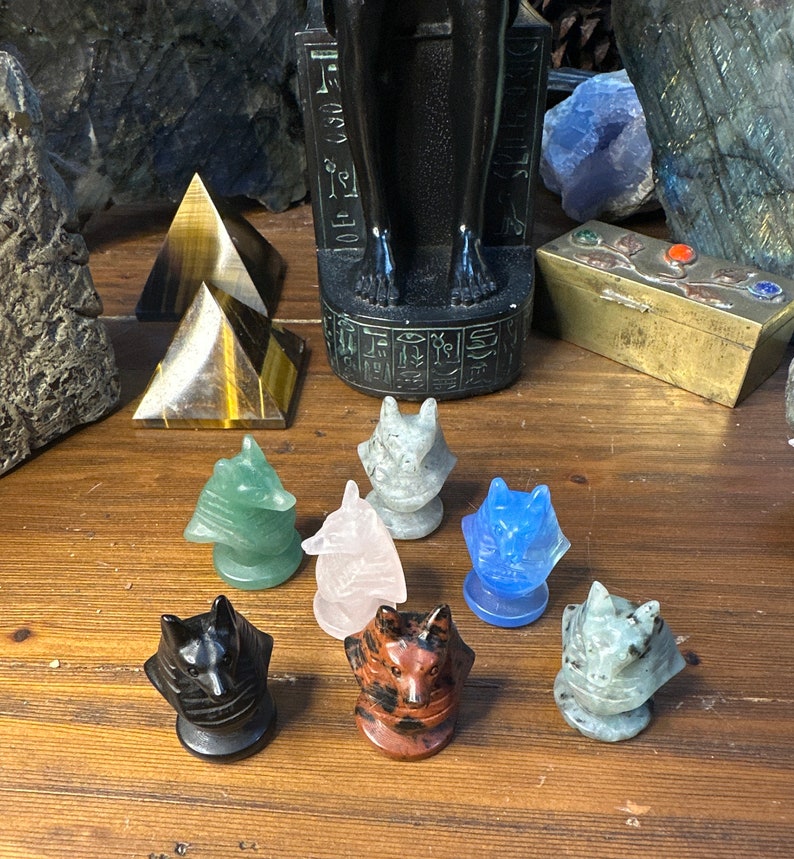 May include: A collection of carved gemstone wolf figurines in various colors, including green, pink, blue, and black. The figurines are displayed on a wooden surface, with a black statue and other crystals in the background.