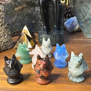May include: A collection of carved stone Anubis figurines in various colors, including black, green, pink, gray, red-brown, and blue. The figurines are displayed on a wooden surface, with other decorative items in the background.