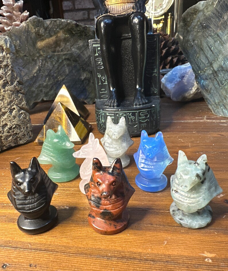 May include: A collection of carved stone Anubis figurines in various colors, including black, green, pink, gray, red-brown, and blue. The figurines are displayed on a wooden surface, with other decorative items in the background.