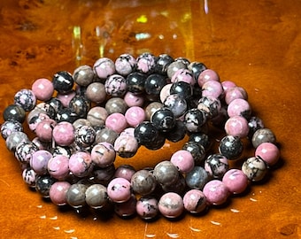 May include: Several beaded bracelets featuring a mix of pink, black, and gray round stones. The bracelets are arranged in a circular pattern on a wooden surface. The stones have a speckled appearance.