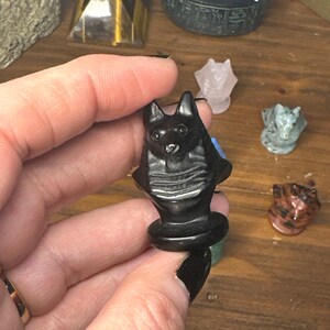 May include: A black carved stone figurine of a cat held in a hand. The cat has pointed ears and a detailed face. Other small animal figurines are visible in the background, including a pink cat and a gray cat.
