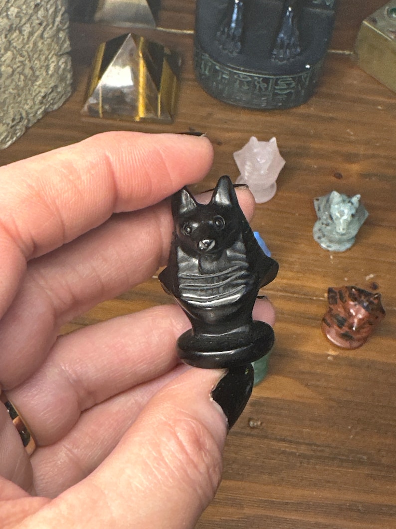 May include: A black carved stone figurine of a cat held in a hand. The cat has pointed ears and a detailed face. Other small animal figurines are visible in the background, including a pink cat and a gray cat.