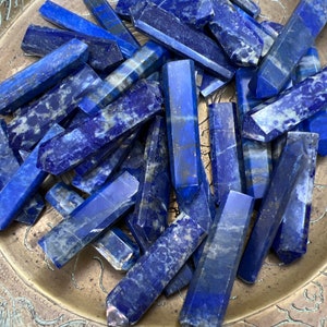 May include: A collection of polished lapis lazuli crystal wands. The wands are a deep blue color with flecks of gold and white. They are hexagonal in shape and vary in length.