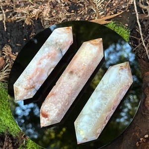 May include: Three polished crystal wands with a mix of white, pink, and brown hues are displayed on a black reflective surface. The crystals are arranged in a row, showcasing their unique patterns and textures. The background features natural elements.