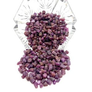May include: A clear, faceted glass bowl filled with small, rough-cut, deep purple gemstones. A pile of the same gemstones spills out from the bowl onto a white surface. The gemstones have a natural, unpolished appearance.