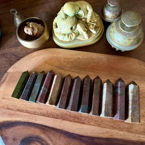 May include: A collection of decorative items on a wooden tray. Includes a small bell, a brass bowl with a textured object, a figurine of two figures, two ornate inkwells, and a row of polished, pointed stone obelisks in shades of green, brown, and copper.