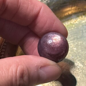 May include: A small, round, purple gemstone held between fingers. The stone has a polished surface with subtle color variations. The background is a silver-colored, decorative tray.