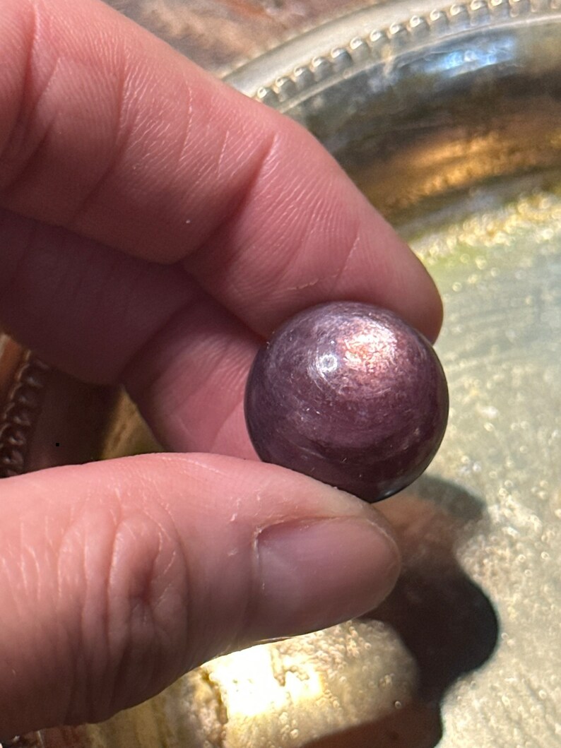 May include: A small, round, purple gemstone held between fingers. The stone has a polished surface with subtle color variations. The background is a silver-colored, decorative tray.