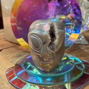 May include: A gray alien head sculpture with a large crack revealing a crystal interior. The sculpture sits on a multi-layered, iridescent, circular stand. A large, iridescent, circular backdrop is behind the sculpture.