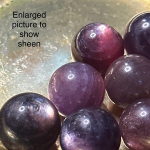 May include: Close-up of several polished, spherical gemstones in shades of purple and dark violet, displayed in a silver-colored dish. The text "Enlarged picture to show sheen" is visible. The gemstones have a reflective sheen.