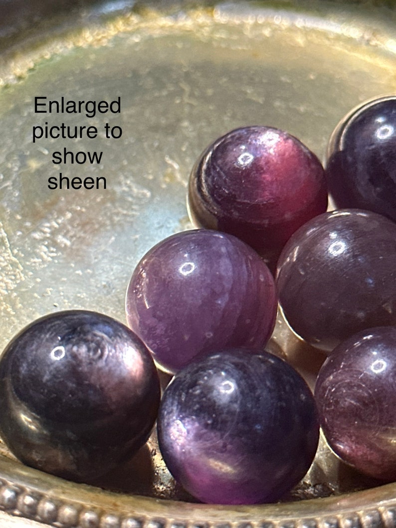 May include: Close-up of several polished, spherical gemstones in shades of purple and dark violet, displayed in a silver-colored dish. The text "Enlarged picture to show sheen" is visible. The gemstones have a reflective sheen.