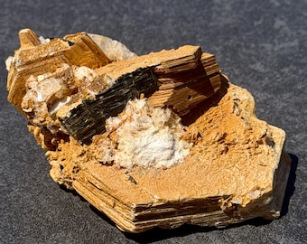 Op de afbeelding: Een natuurlijk mineraal specimen met gelaagde, goudbruine en zwarte kristallen met witte kwartsinsluitsels. De ruwe steen heeft een textuur, gepresenteerd tegen een donkere achtergrond.