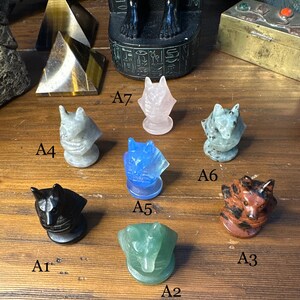 May include: A collection of carved stone wolf figurines in various colors, including black, blue, green, pink, and brown. Each wolf head is mounted on a circular base. Other stone objects are in the background.