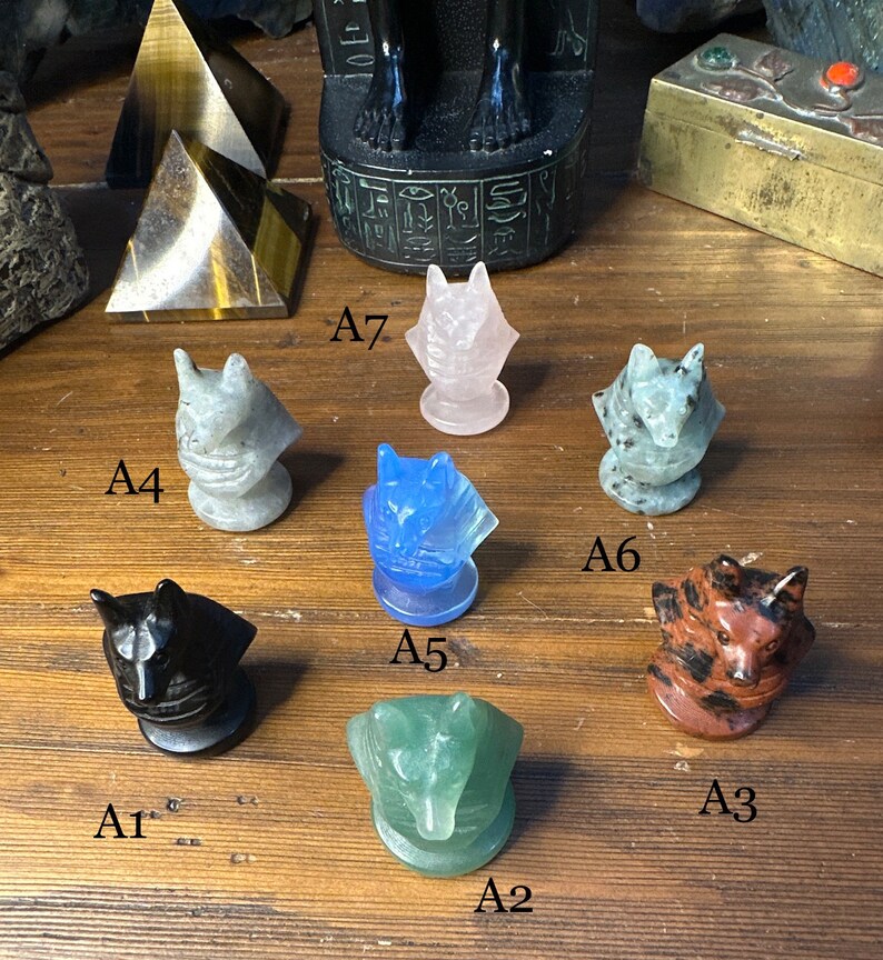 May include: A collection of carved stone wolf figurines in various colors, including black, blue, green, pink, and brown. Each wolf head is mounted on a circular base. Other stone objects are in the background.