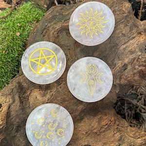 May include: Four round, light pink crystal stones with gold-colored symbols. The symbols include a sun, a pentagram, a goddess symbol, and moons with stars. The stones are displayed on a piece of wood.