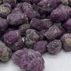 May include: A collection of rough, uncut gemstones in shades of deep purple and gray. The stones have a textured, uneven surface, and vary in size. The image is a close-up, showing the details of the natural crystals.