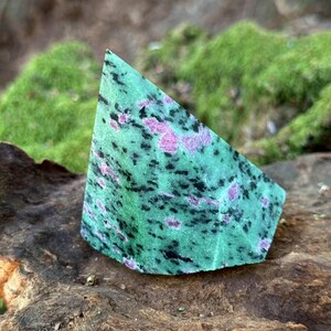 May include: A triangular-shaped, polished gemstone with a vibrant green base color. The stone is speckled with black and pinkish-red inclusions. It rests on a piece of weathered wood, with a blurred green moss background.