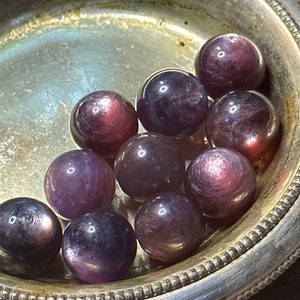 May include: A collection of polished, spherical gemstones in shades of purple and burgundy, displayed in a silver dish. The gemstones have a reflective, iridescent quality, with subtle variations in color and texture. The dish has a decorative, ridged edge.