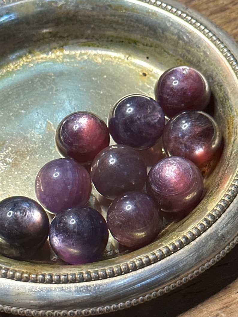 May include: A collection of polished, spherical gemstones in shades of purple and burgundy, displayed in a silver dish. The gemstones have a reflective, iridescent quality, with subtle variations in color and texture. The dish has a decorative, ridged edge.